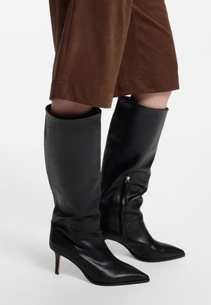 Personne portant des bottes en cuir noires montantes jusqu'au genou, à bout pointu, avec de petits talons et un pantalon marron court et large.
