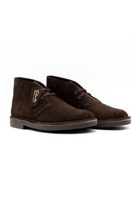 Botas de tobillo de ante marrón con punta redonda, cordones y suela de goma cosida. Presentan una etiqueta que indica "Clarks Desert Boot."