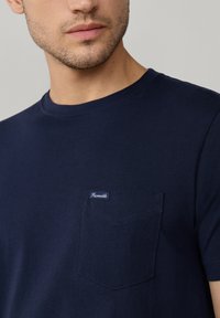 Granatowa bawełniana koszulka z okrągłym dekoltem, krótkimi rękawami i kieszenią na piersi, ozdobiona małą metką marki w białym i niebieskim kolorze.