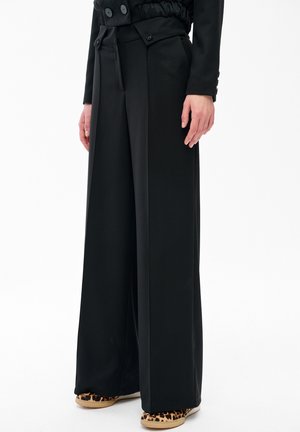 Osoba ubrana w czarne spodnie o szerokich nogawkach z guzikiem na pasku oraz w buty wsuwane w panterkę, stojąca na tle białego tła.