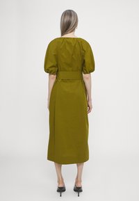 Olivově zelené midi šaty s nabíranými rukávy, opaskem v pase a hladkou texturou. Nosené s černými vysokými podpatky. Pohled zezadu.