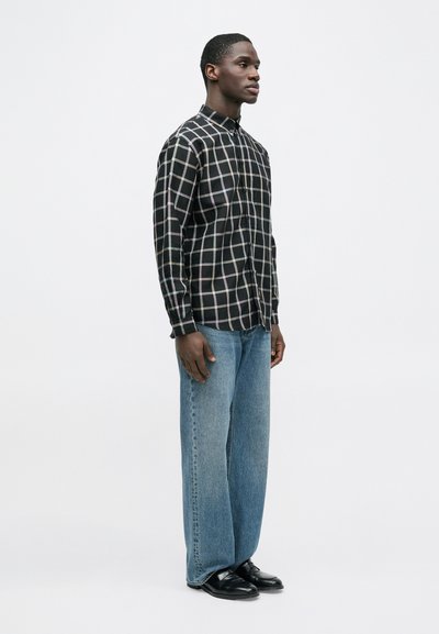 Μαύρο πουκάμισο με καρό σχέδιο και κουμπιά, με μακριά μανίκια, σε συνδυασμό με ανοιχτό μπλε τζιν με φαρδιά παντελόνια και μαύρα παπούτσια. Απλός και μοντέρνος σχεδιασμός.