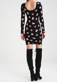 Robe noire à manches longues avec un motif floral rouge et blanc ; design ajusté avec un dos dénudé et une longueur mi-genou. Portée avec de hautes bottes en daim noires.