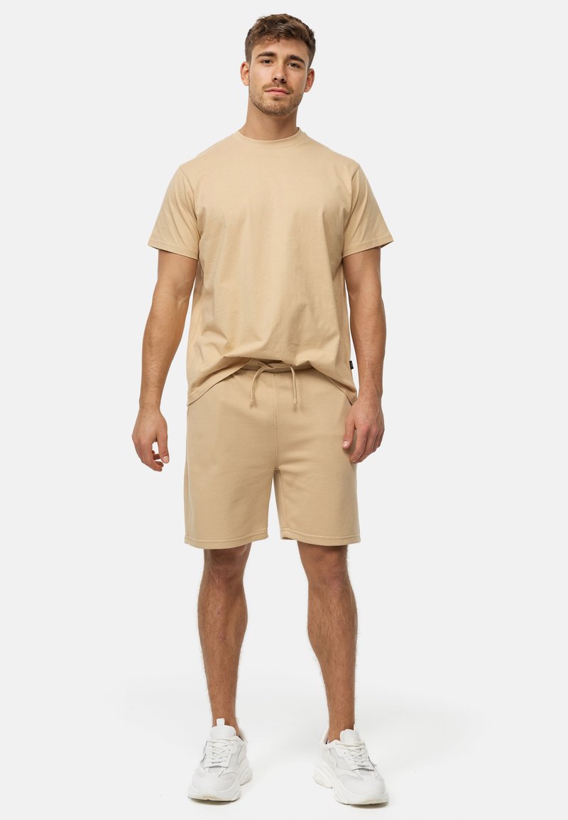 Ung mand står oprejst iført beige T-shirt og matchende shorts med hvide sneakers mod ensfarvet baggrund.