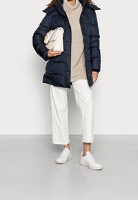 Granatowa puchowa kurtka z wysokim kołnierzem, beżowy sweter z dzianiny, białe spodenki do kostek i białe sneakersy. Beżowa torebka z łańcuszkiem.