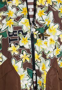 Blumengemusterte gesteppte Jacke mit braunem Grund, die gelbe und weiße Blumen zeigt. Vorderer Reißverschluss, schwarzer Reißverschluss und kontrastierende braune Stoffakzente.