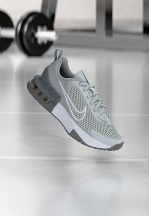 Grauer Nike Air Max Sneaker mit weißem Swoosh, Mesh-Design und sichtbarer Luftpolstersohle, schwebend in einem Fitnessstudio mit unscharfen Gewichten im Hintergrund.