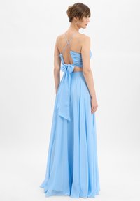 Frau, die ein bodenlanges, hellblaues Kleid mit gerafftem Mieder, dünnen überkreuzten Trägern, offener Rückseite und einer großen Schleife am unteren Rücken trägt.