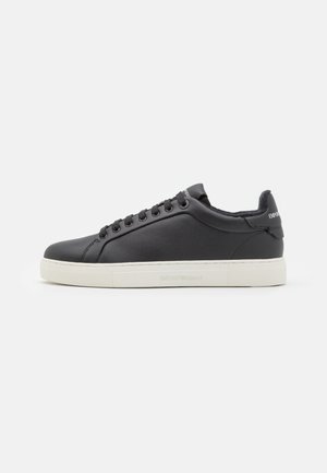 Baskets en cuir noir avec un bout rond, semelle en caoutchouc blanc plat et accents texturés. Présente des lacets plats et des détails de marque sur le talon.