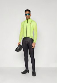 Dzeltena riteņbraukšanas jaka ar garām piedurknēm, melnas legingi un melni riteņbraukšanas apavi; kopā ar melnu ķiveri un stilīgām saulesbrillēm.