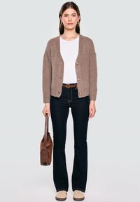 Hellbrauner Strickcardigan mit Knöpfen, über einem weißen Hemd getragen, kombiniert mit dunkelblauen Schlagjeans und beigen Turnschuhen, während eine braune Tasche gehalten wird.