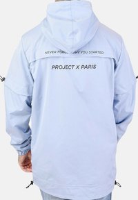 Giacca a vento blu chiaro con cappuccio, polsini elastici e orlo con cordino. Presenta testo nero in grassetto sul retro: "NON DIMENTICARE MAI PERCHÉ HAI INIZIATO" e "PROJECT X PARIS."