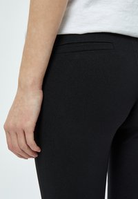 Pantaloni neri aderenti con una texture liscia, dotati di una piccola tasca posteriore e un design elegante. Braccia visibili poggiano sul tessuto.
