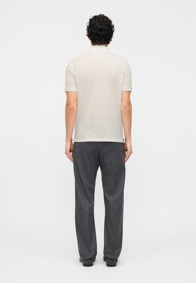 Άνδρας με σγουρά σκούρα μαλλιά στέκεται γυρισμένος προς τα πίσω, φορώντας κοντομάνικο ανοιχτό μπεζ πόλο και φαρδιά σκούρα γκρι παντελόνια.