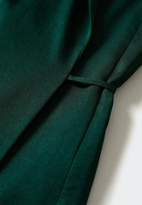 Tissu vert foncé à texture lisse, avec un détail noué au niveau de la manche. Le design présente une forme drapée et des plis doux.