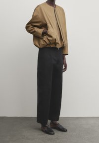 Veste bomber beige avec fermeture éclair, taille élastique et grandes poches, associée à un pantalon large noir et des chaussures plates noires.