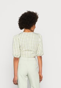 Blusa curta de gingham verde e branco com mangas bufantes; apresenta punhos elásticos e um simples decote redondo. Usada com calças de cor clara.