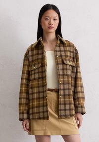 Bruin en tan geruit flanellen shirt met lange mouwen, twee voorzakken en gele knopen, gedragen over een beige top en rok.