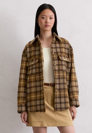 Braun und beige kariertes Flanellhemd mit langen Ärmeln, zwei vorderen Taschen und gelben Knöpfen, getragen über einem beigen Oberteil und einem Rock.