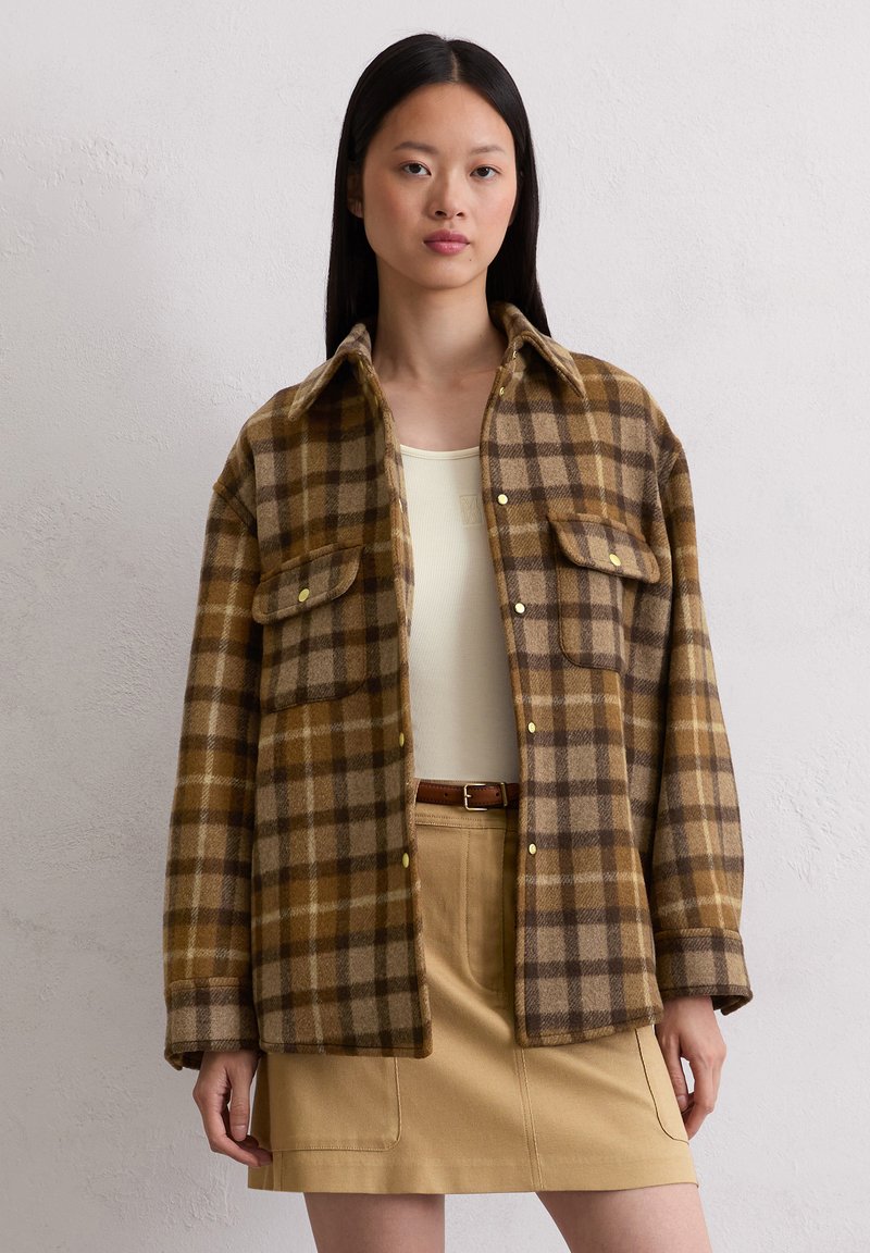 Bruin en tan geruit flanellen shirt met lange mouwen, twee voorzakken en gele knopen, gedragen over een beige top en rok.