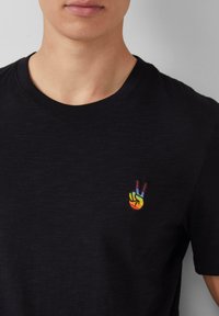 Man die een zwart T-shirt draagt met een kleine regenboogkleurige vredeshand-icoon geborduurd op de linkerkant van de borst.