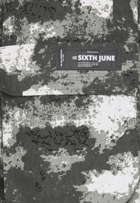 Kamouflage-mönstrad tyg i grått och vitt, med ett sytt etikett som står "SIXTH JUNE." Texturerad yta med varierande nyanser och fläckar.
