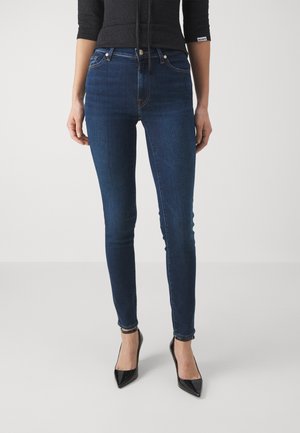Jean skinny bleu foncé avec une texture en denim lisse, comprenant cinq poches, un matériel en métal argenté et un discret détail de couture le long des coutures.