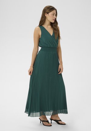 Femme aux longs cheveux bruns portant une robe maxi sans manches plissée de couleur vert foncé et des sandales à talons noires, tournée vers la gauche sur un fond uni.