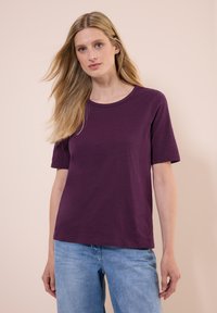 T-shirt viola a maniche corte realizzato in tessuto morbido, con scollatura rotonda e vestibilità comoda. Abbinato a jeans azzurri chiari.