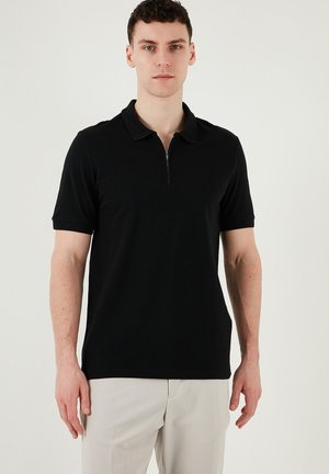 Black polo shirt with a zippered collar, short sleeves, and a smooth texture. Worn with light-colored trousers, minimalist design.