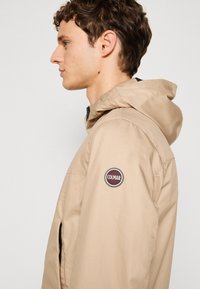 Beige hooded jacket made of lightweight material, featuring side zip pockets and a round Colmar logo patch on the left arm.