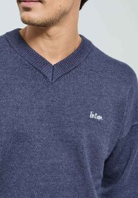 Pull en tricot bleu marine avec un design à col en V, doté de bords ribbed et d'un petit logo blanc sur la poitrine. Texture lisse.