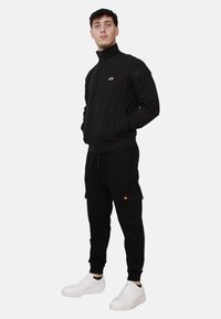 Giacca nera con zip e collo alto, abbinata a pantaloni jogger neri con tasche cargo. Il modello indossa sneakers bianche. Design semplice e sportivo.