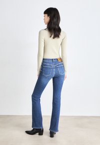 Jeans flare in denim blu con vita alta, cinque tasche e un'etichetta in pelle beige sul retro. Indossati con un top a maniche lunghe a coste di un beige chiaro.