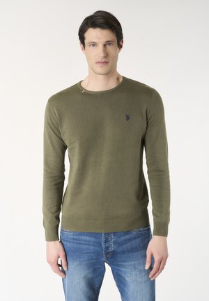 Giovane uomo con capelli scuri che indossa un maglione verde oliva a maniche lunghe e jeans blu, in piedi davanti a uno sfondo chiaro e uniforme.