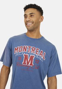 Blauwe katoenen t-shirt met ronde hals. Kenmerkt zich door de tekst "MONTREAL" in vet rood en wit, met daaronder "1972 Running Club".