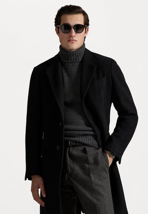 Musta villast mantel struktuurse disainiga, kantud tumeda halli ribitev turtlenecki ja hallide pükste peal; mustad prillid ja kell on kaasas.