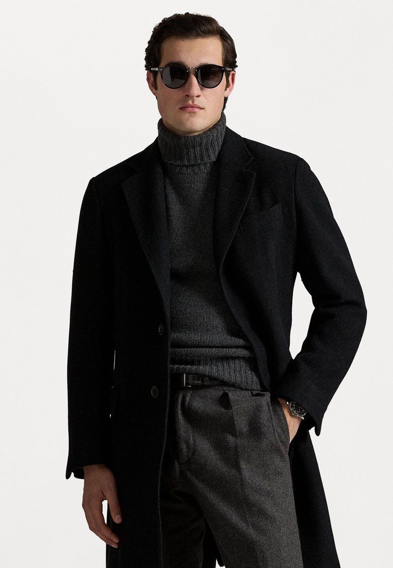 Black wool coat with a structured design, worn over a dark gray ribbed turtleneck and gray trousers; black sunglasses and watch included.