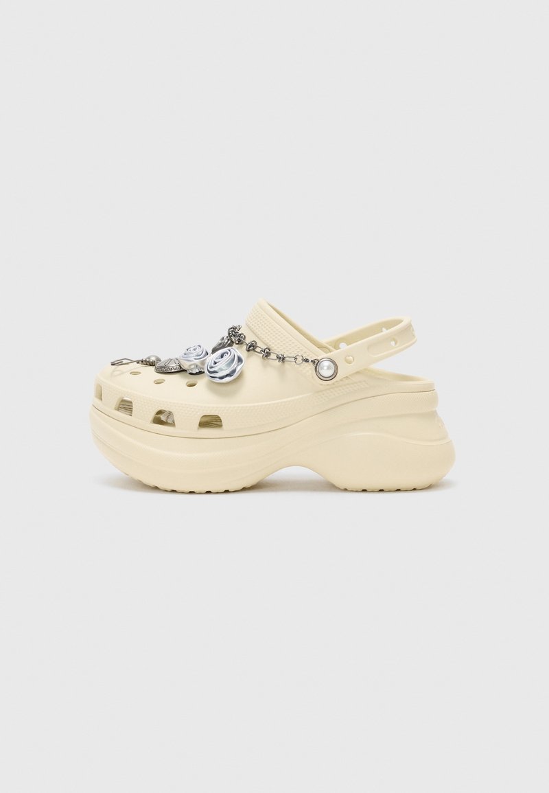 Sabot à plateforme beige avec une bride arrière, des trous de ventilation et une chaîne argentée décorative ornée de fleurs et de breloques à l’avant.