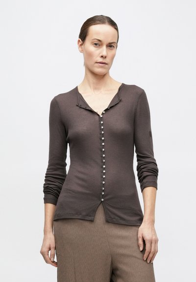 Hátra kötött hajjal rendelkező nő, szűk, gombos sötétbarna kardigánt és laza világosbarna nadrágot viselve, egy egyszínű világos háttér előtt áll.