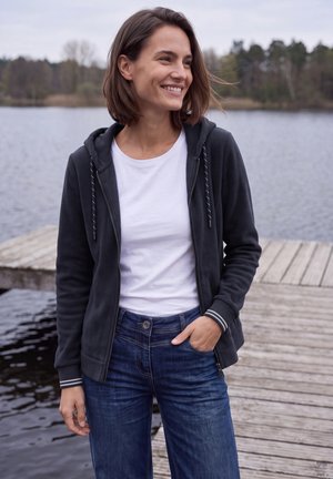 Mujer sonriendo, de pie en un muelle de madera junto al lago, vistiendo una camiseta blanca, sudadera oscura con cremallera y vaqueros azules, con árboles al fondo.