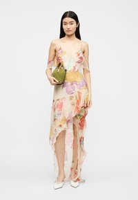 Γυναίκα που φοράει διαφανές φόρεμα με floral μοτίβο σε στυλ υψηλό-χαμηλό, κρατώντας μια μικρή πράσινη τσάντα, συνδυασμένο με λευκές μυτερές γόβες, στέκεται σε ουδέτερο φόντο.