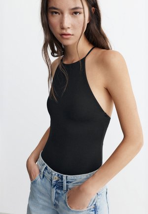 Jeune femme aux longs cheveux foncés portant un haut noir sans manches et un jean en denim bleu clair, les mains dans les poches.