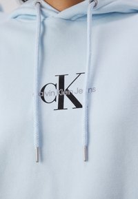 Sudadera azul claro con cordones, que cuenta con un logo impreso en negro "CK" y el texto "calvin Klein Jeans" debajo en gris. Textura de tela suave.