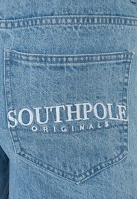 Tejido de mezclilla azul claro con un bolsillo trasero que presenta el texto bordado "SOUTHPOLE" y "ORIGINALS". Costuras visibles y detalles de textura.