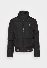 Svart puffjacka med hög krage, zipstängning och vita logodetaljer. Har quiltade sektioner och elastiska muddar och fåll.