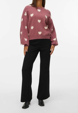 Femme portant un pull mauve avec des motifs de cœurs blancs, un pantalon noir large et des bottines à talons noires sur fond blanc.