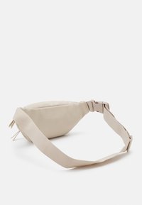 Béžová ledvinka z hladkého materiálu, která má zakřivený tvar, nastavitelný popruh a zip s ozdobnými střapci.