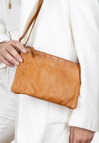 Borsa a tracolla in pelle color tan con finitura testurizzata, chiusura con zip e una sottile tracolla, tenuta in mano contro un outfit bianco.