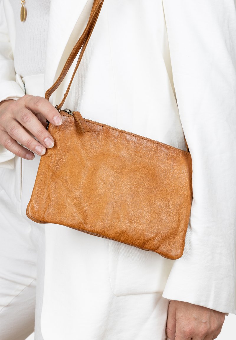 Borsa a tracolla in pelle color tan con finitura testurizzata, chiusura con zip e una sottile tracolla, tenuta in mano contro un outfit bianco.